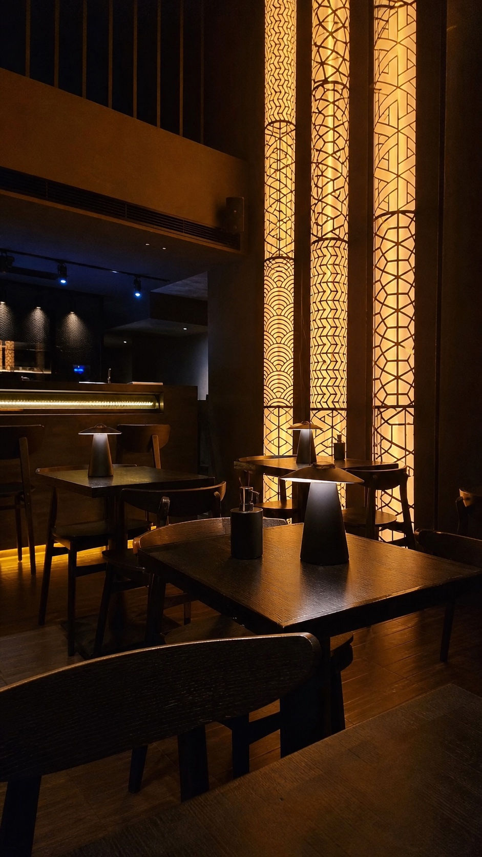 Study of the Koyasan dining room from a seat at the table: patterned columns of amber light beside dark timber tables and lamps.