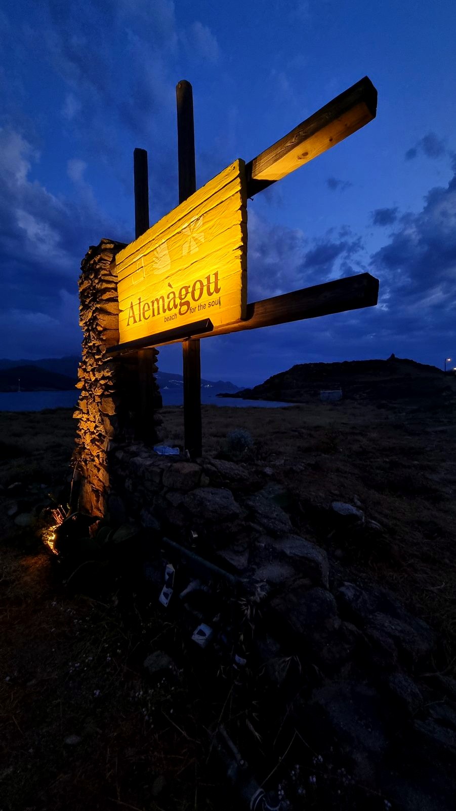 The timber Alemàgou sign lit warm against a blue hour sky beside a dry stone pillar.