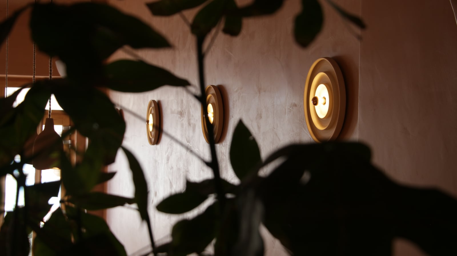 Three round sconces glowing along a plaster wall at Bouboulo, seen through the dark silhouettes of leaves.