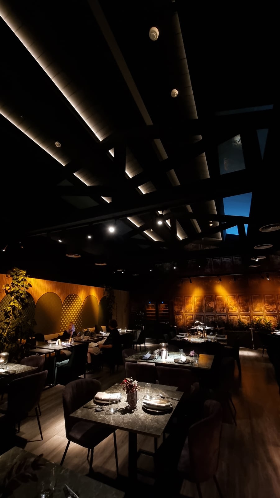 The Itameshi dining room at night: lines of light along the dark ceiling beams, skylights above and a wall of painted panels lit warm at the far end.