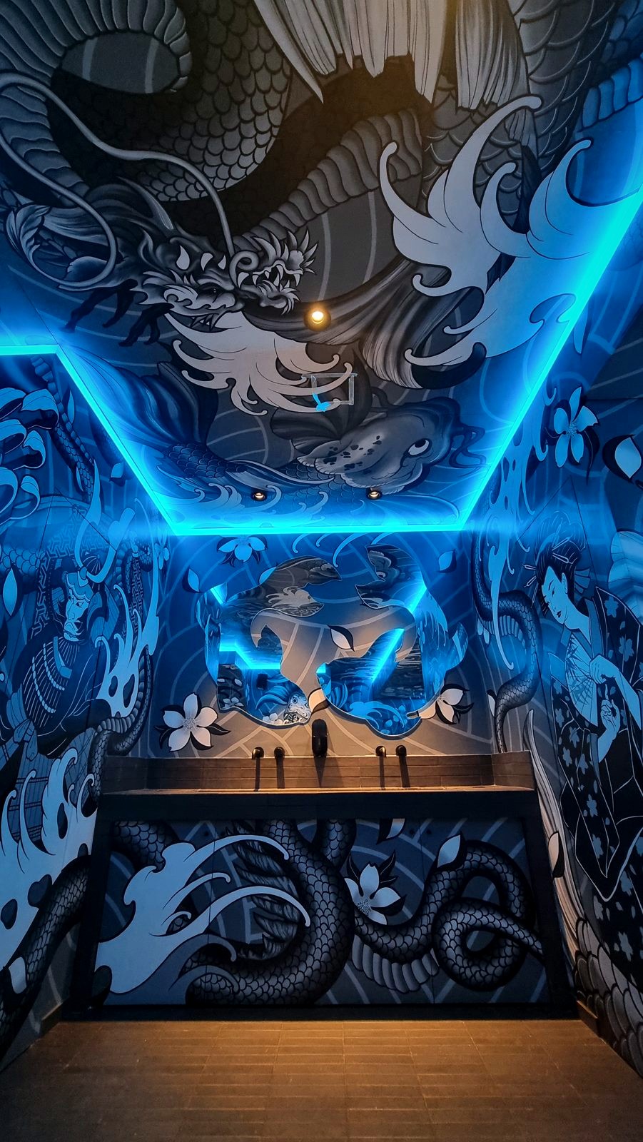 A Koyasan washroom wrapped in a black and white dragon mural, lit by a blue cove above a long basin.