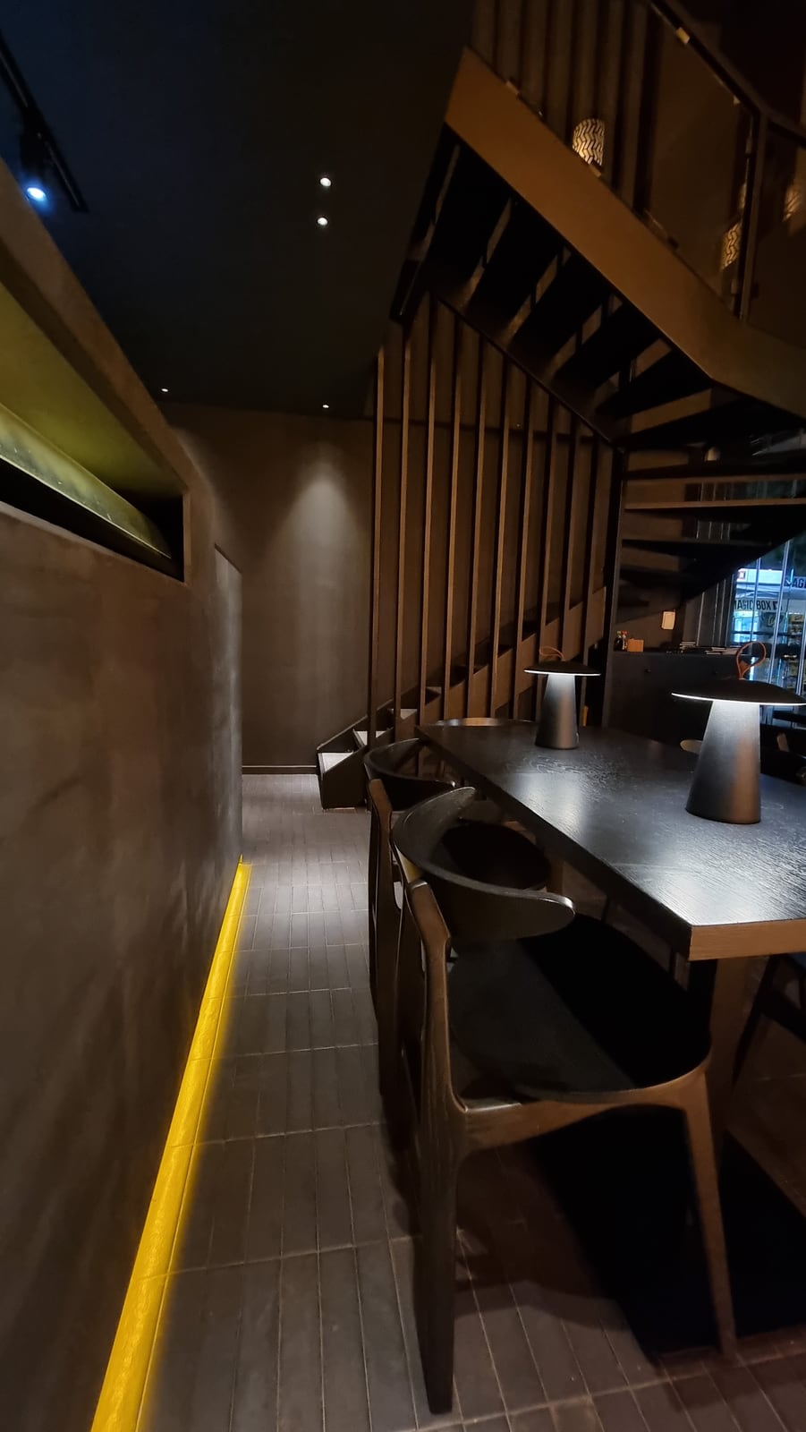 A long dark table with lamps beneath the slatted timber stair at Koyasan, a line of gold light along the foot of the wall.