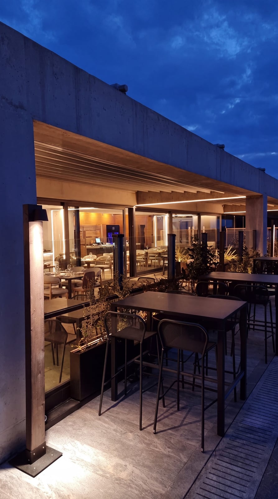 High tables on the Nyn Esti rooftop terrace at dusk, warm light along the underside of the canopy and the glazed dining room behind.
