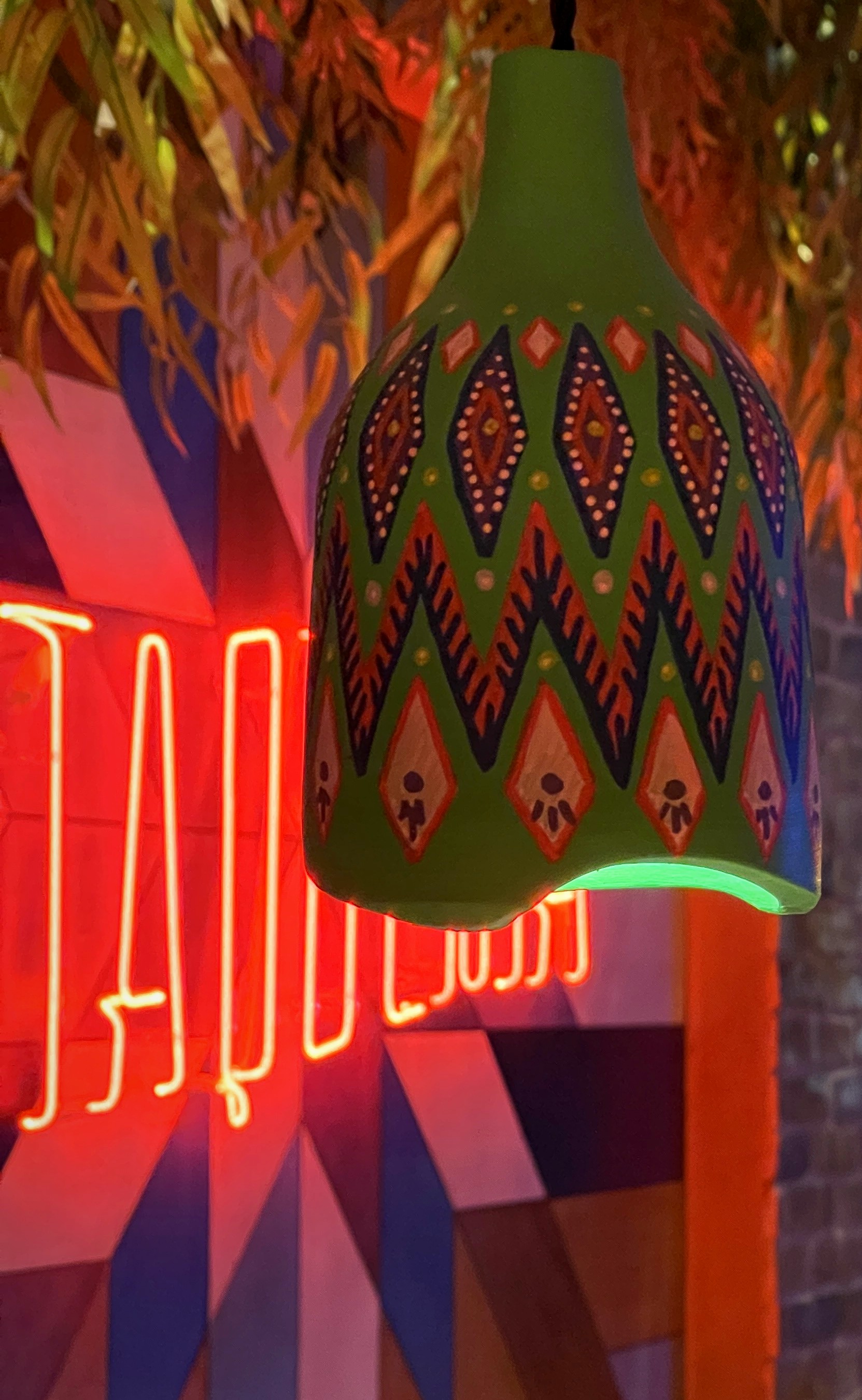 A green hand painted pendant glowing at its rim in front of red neon and a patterned wall at Panchito.
