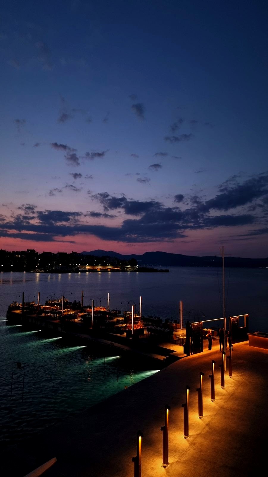 The Raval jetty at dusk, traced by low bollard lights with a soft glow beneath the water.