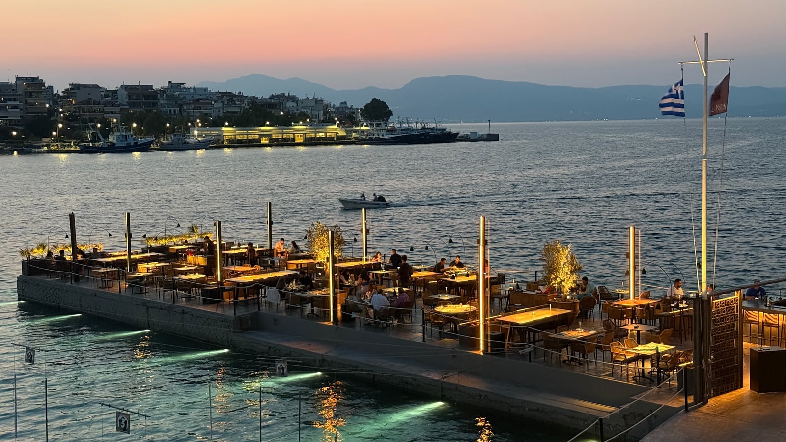 The Raval terrace from above at sunset, tables set along the jetty, low lights at its edge and the town and hills across the water.