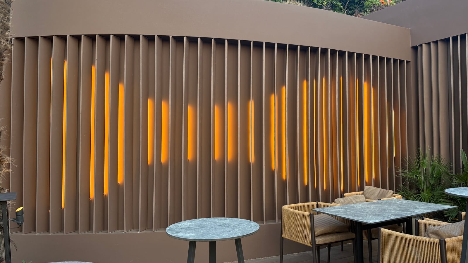 Warm lines of light between the vertical slats of a curved wall at Raval, marble tables and woven chairs before it.