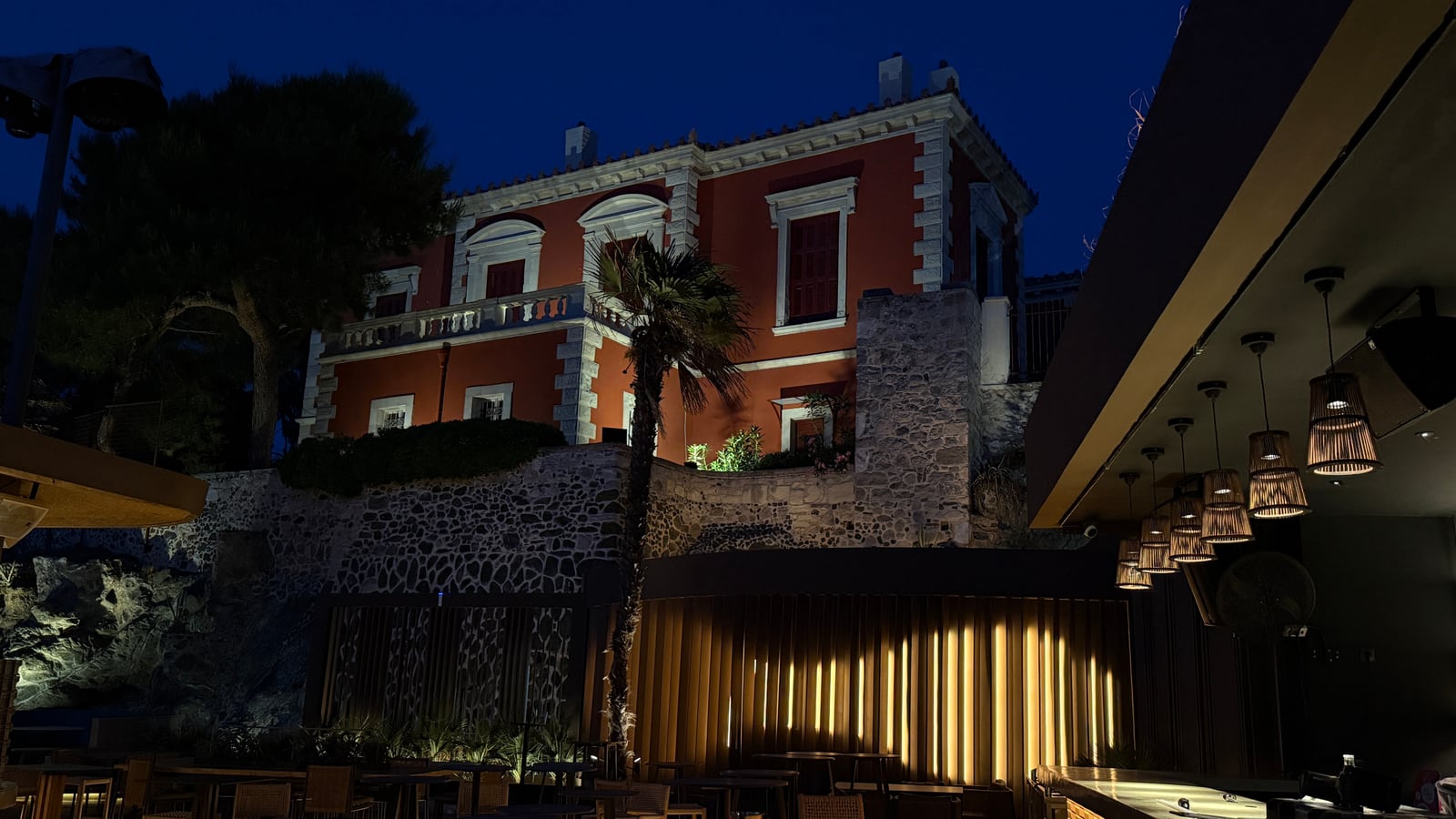 A red villa above the Raval terrace at blue hour, a palm before it, rattan pendants under the canopy and warm light between the slats of the fence.