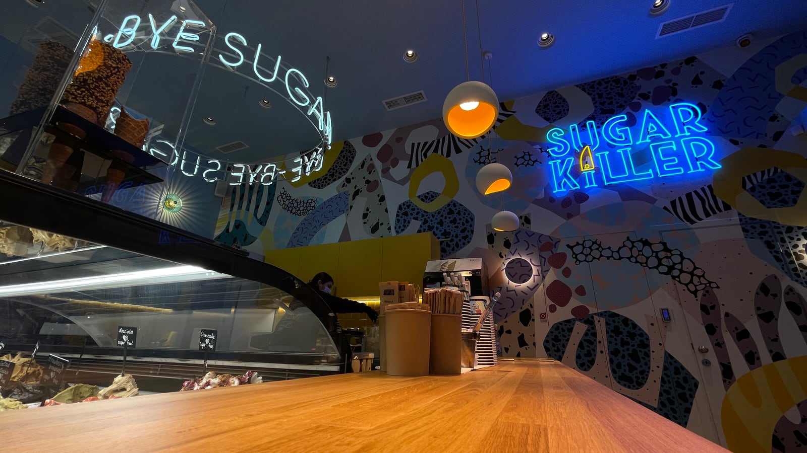 Sugar café's timber counter under orange lit pendants, a ring of neon letters over the glass display and a blue neon sign on the patterned mural.