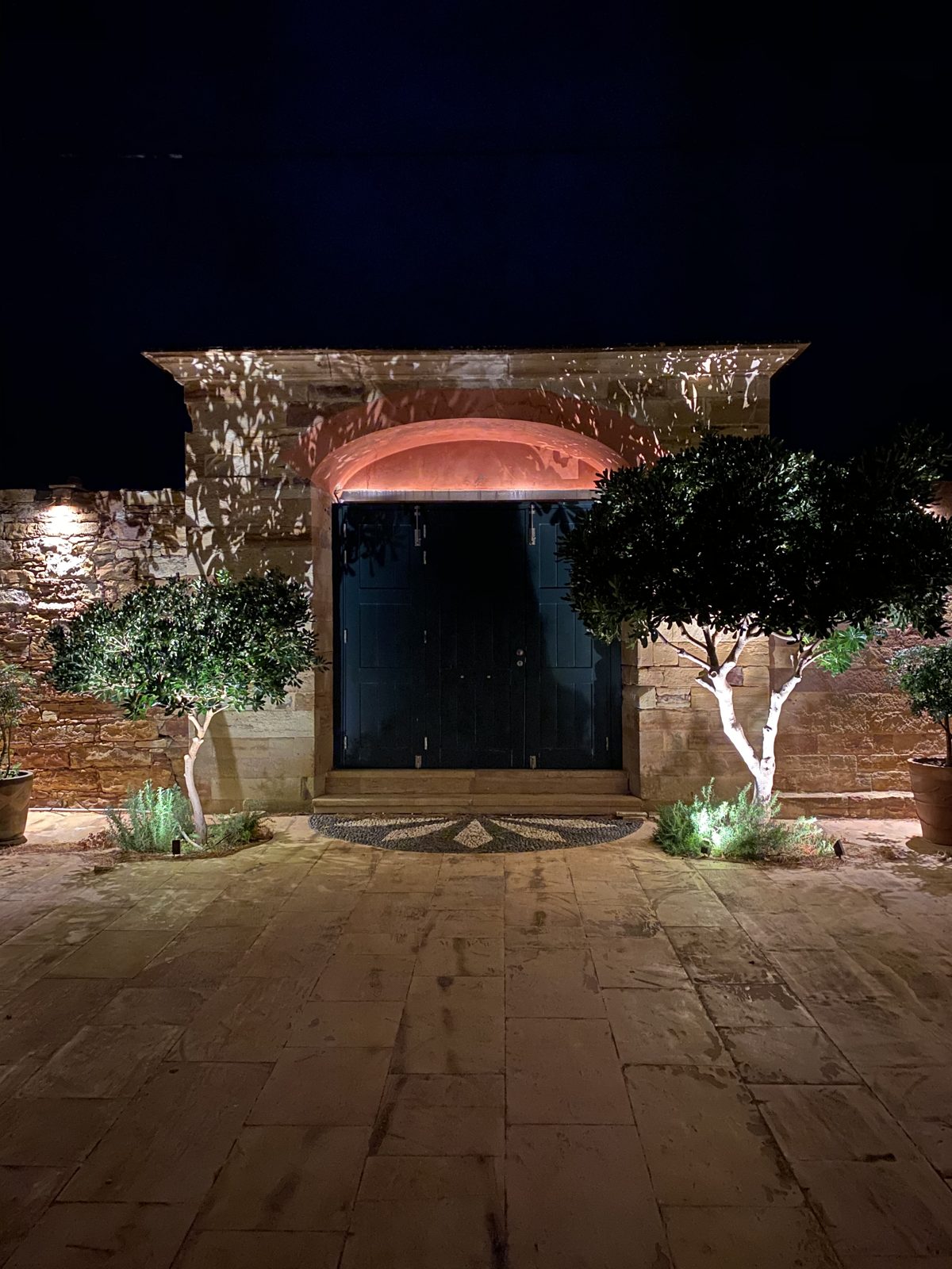 The villa's timber gate under a softly lit arch, with a mosaic threshold and uplit olive trees before it.