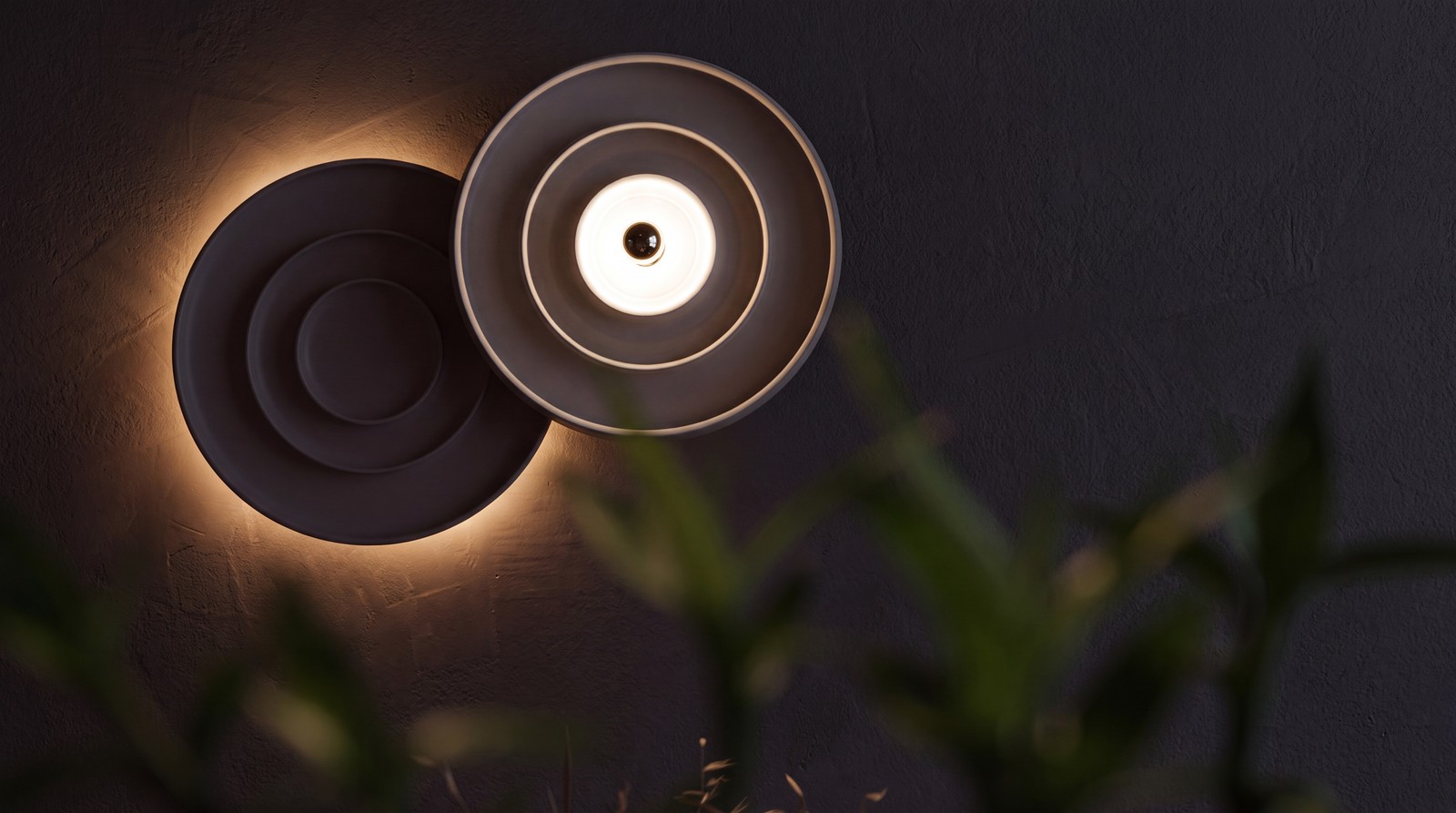 Two round wall lights, one lit, casting a warm halo across a dark wall